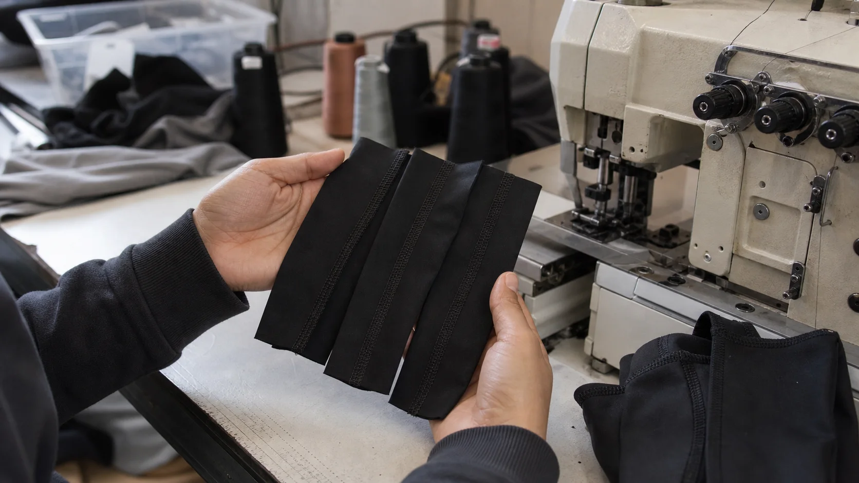 Hands comparing black activewear seam samples beside an industrial coverstitch machine
